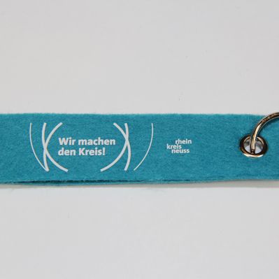 Türkisfarbener Schlüsselanhänger aus Filz mit dem Aufdruck „Wir machen den Kreis“ und „rhein kreis neuss“ jeweils in weiß. Der Schlüsselanhänger hat eine Öse und einen Schlüsselring. 