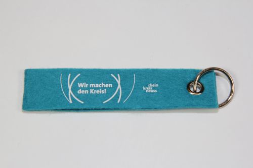 Türkisfarbener Schlüsselanhänger aus Filz mit dem Aufdruck „Wir machen den Kreis“ und „rhein kreis neuss“ jeweils in weiß. Der Schlüsselanhänger hat eine Öse und einen Schlüsselring. 